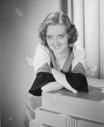 Bette Davis - Bette Davis Leaning on a Table with Arms Crossed in Two Tone Long Sleeve Shirt and Velvet Skirt